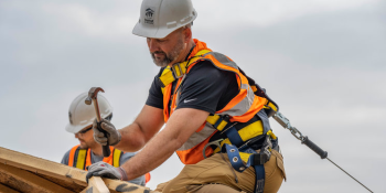 Man in construction gear and hard hat hammers nails on top of a roof