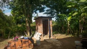 Photo of a small, brick structure, a latrine, near green trees.
