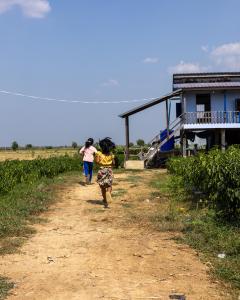 Two young girls running on a dirt path toward a blue house on stilts.