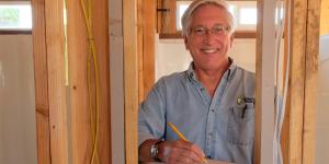 Cliff in a blue shirt, holding a pencil to paper as he stands in a partially constructed house.