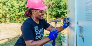 A woman in a pink hardhat and homeowner T-shirt smiles as she hammers.