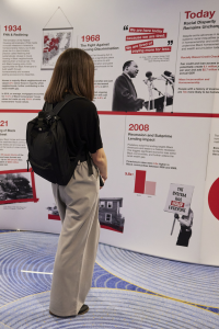 A woman is seen from behind as she reads a large exhibit display with a timeline including the years 1934, 1968, 2008, and today. The display also features an image of MLK Jr with quote "We are here today because we are tired. We are tired of paying more for less."