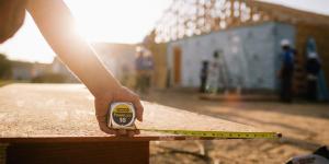 Bright sunset shines in the background with arm holding measuring tape on house construction site in foreground.