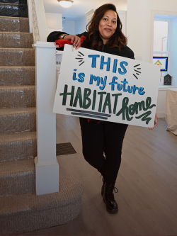 Woman standing next to stairs inside a house, holding a poster that reads "This is my future Habitat home!"