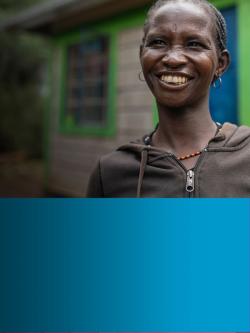 Portrait of a Kenyan woman smiling at the camera in front of a grey brick house with colorful green trim around the windows and a blue door.