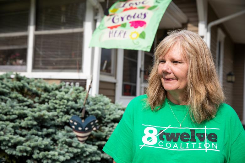 Women in a T-shirt that says "8 twelve" stands in front of a home.