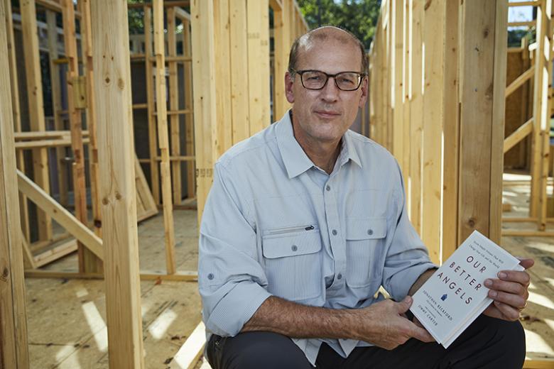 Jonathan Reckford sitting in wood framed house holding Our Better Angels book.