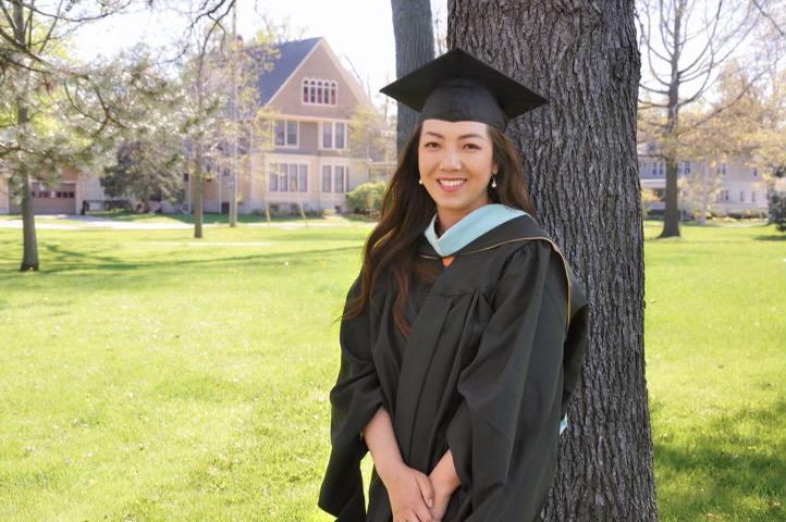 Ai posing by a tree in her graduation cap and gown.