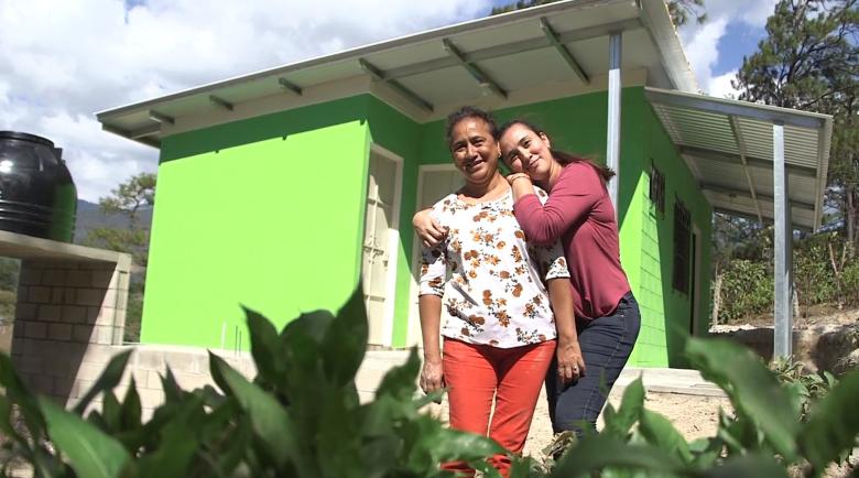 Two women hugging in front of their bright green home in El Salvador