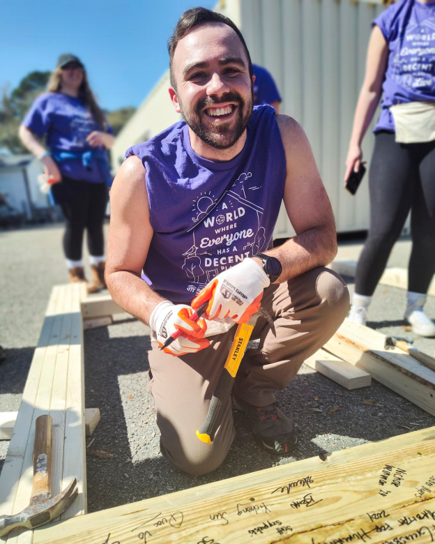 Collegiate Challenge volunteer in purple Habitat shirt smiles as he signs wood framing on a build site.