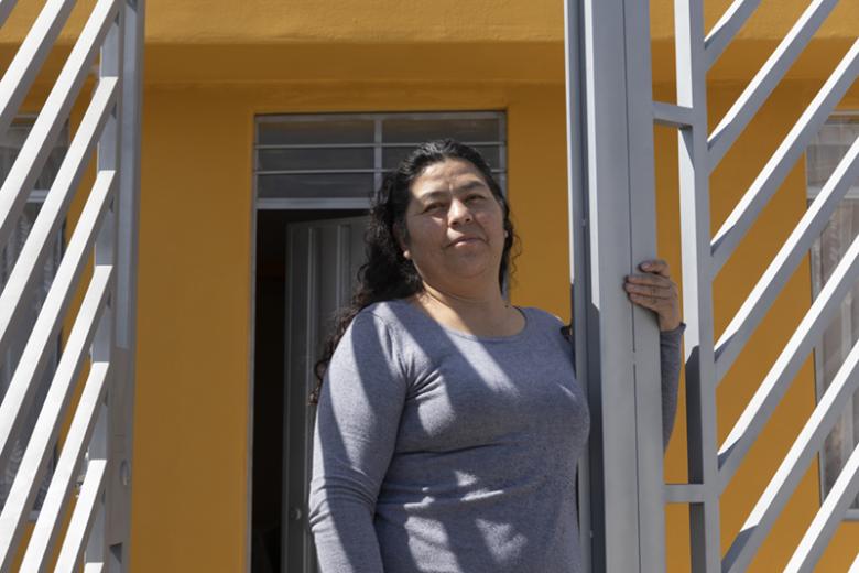 Peruvian woman stands looking at the camera in the gateway of a tall metal fence in front of her bright yellow home.