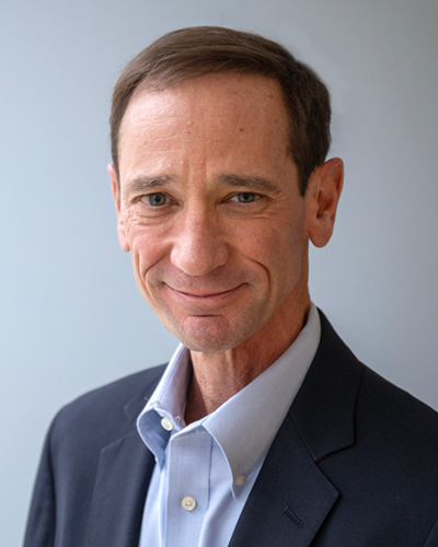 Headshot portrait of Craig Molyneaux smiling at the camera in a black suit and light blue collared shirt.