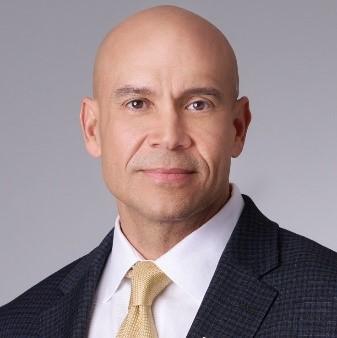 Headshot portrait of Tim Phillips in a dark suit with white shirt and gold tie.