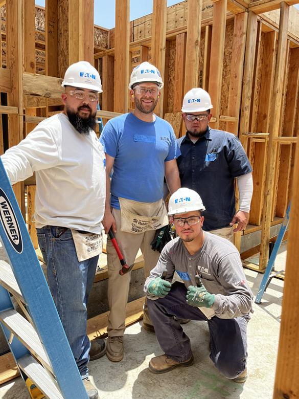 Four men wearing hardhats that say "Eaton" pose together in the wood framing of a Habitat house construction site.