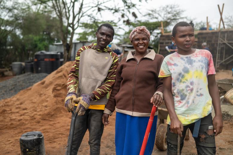 A woman and two young boys stand in front of a construction site holding shovels